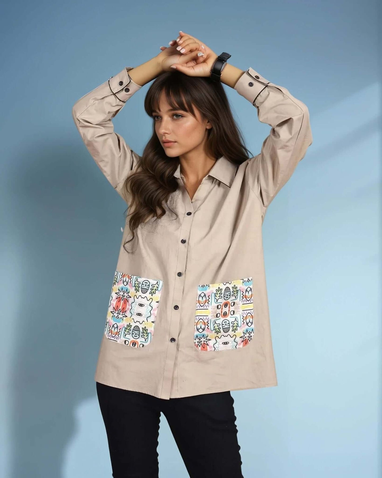 A model poses in a beige oversized button-down shirt featuring bold black buttons and colorful tribal-inspired printed pockets. The relaxed fit and statement pocket design add a touch of bohemian charm, making it a stylish choice for casual wear.
