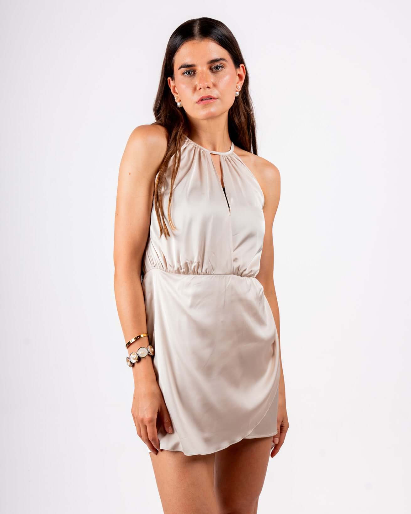 woman wearing a sleeveless, satin mini dress in a soft champagne color. The dress has a high halter neckline and a relaxed, draped fit, creating an elegant and chic look.