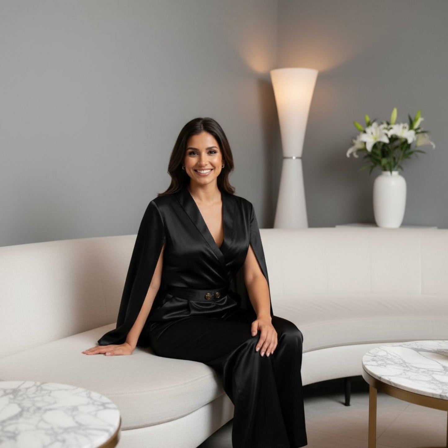 Indian model wearing the Midnight Luxe Cape Jumpsuit by House of Majisha a satin black jumpsuit with cape sleeves and gold belt detail, styled in a modern interior with white dΓ©cor.