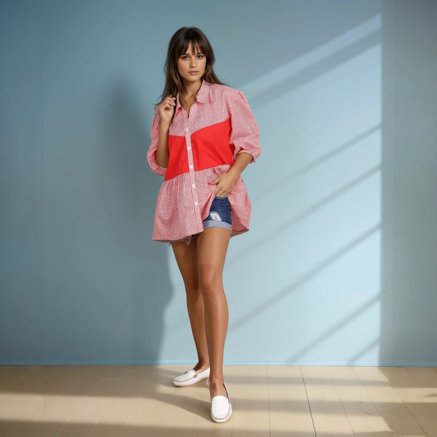 A model wearing an oversized red and white gingham shirt with a bold red panel, paired with denim shorts and white loafers, standing confidently against a pastel blue backdrop. The casual yet trendy outfit exudes effortless summer chic.