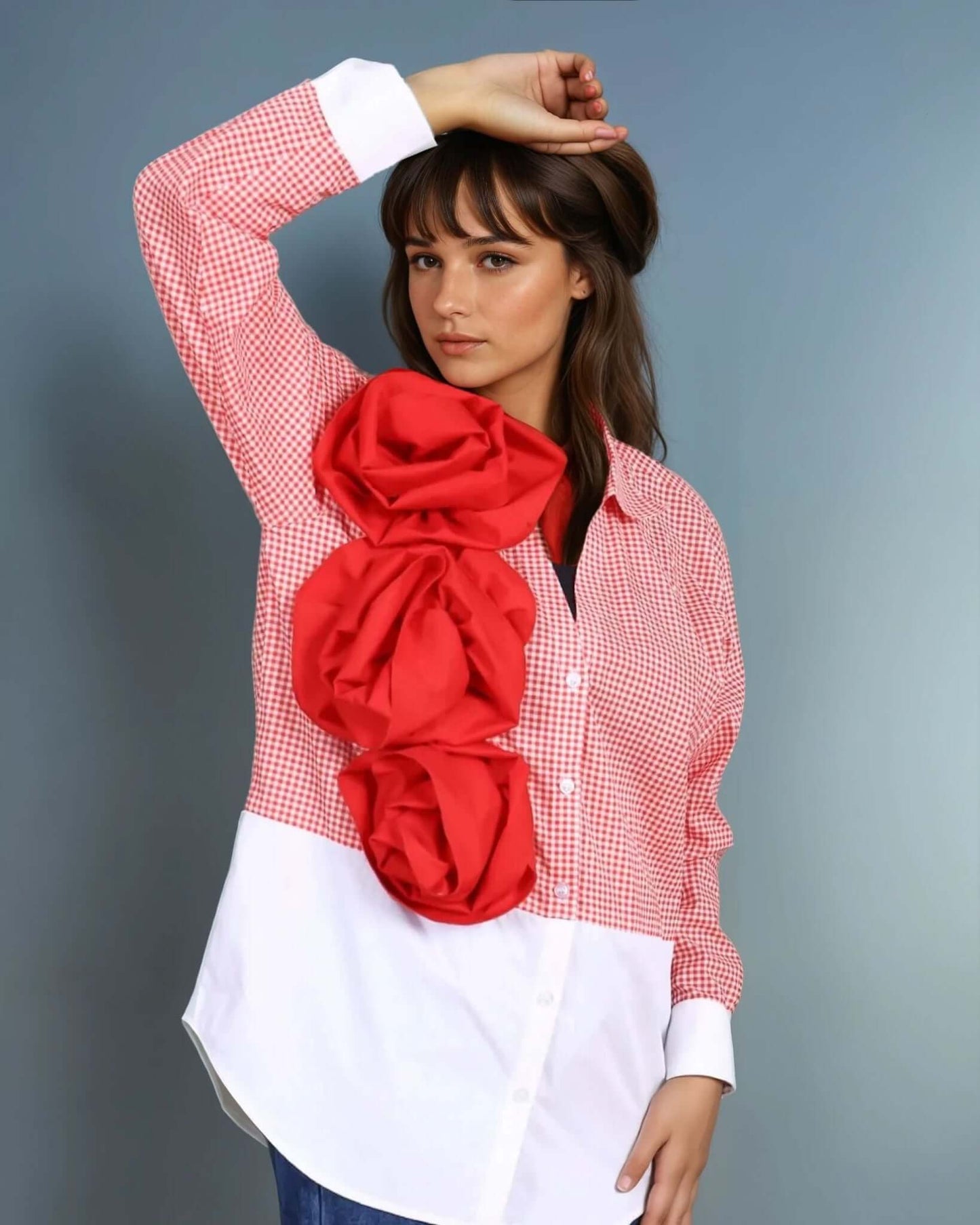 A model poses in a red and white gingham oversized shirt with a bold three-dimensional red floral applique from House Of Majisha, paired with wide-leg blue denim jeans and metallic heels. The structured yet playful design makes this shirt a standout statement piece, perfect for contemporary and fashion-forward styling.
