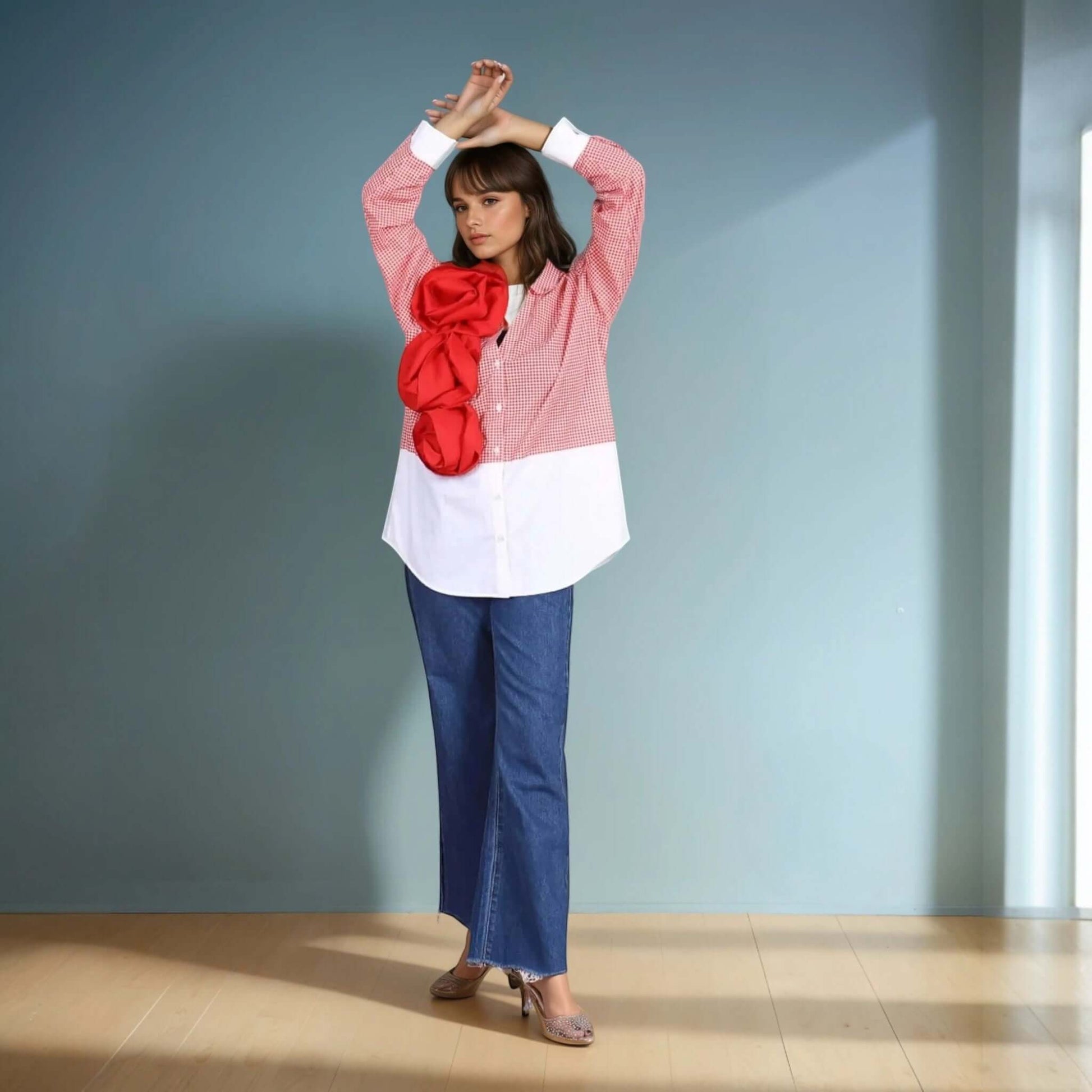 A model poses in a red and white gingham oversized shirt with a bold three-dimensional red floral applique from House Of Majisha, paired with wide-leg blue denim jeans and metallic heels. The structured yet playful design makes this shirt a standout statement piece, perfect for contemporary and fashion-forward styling.