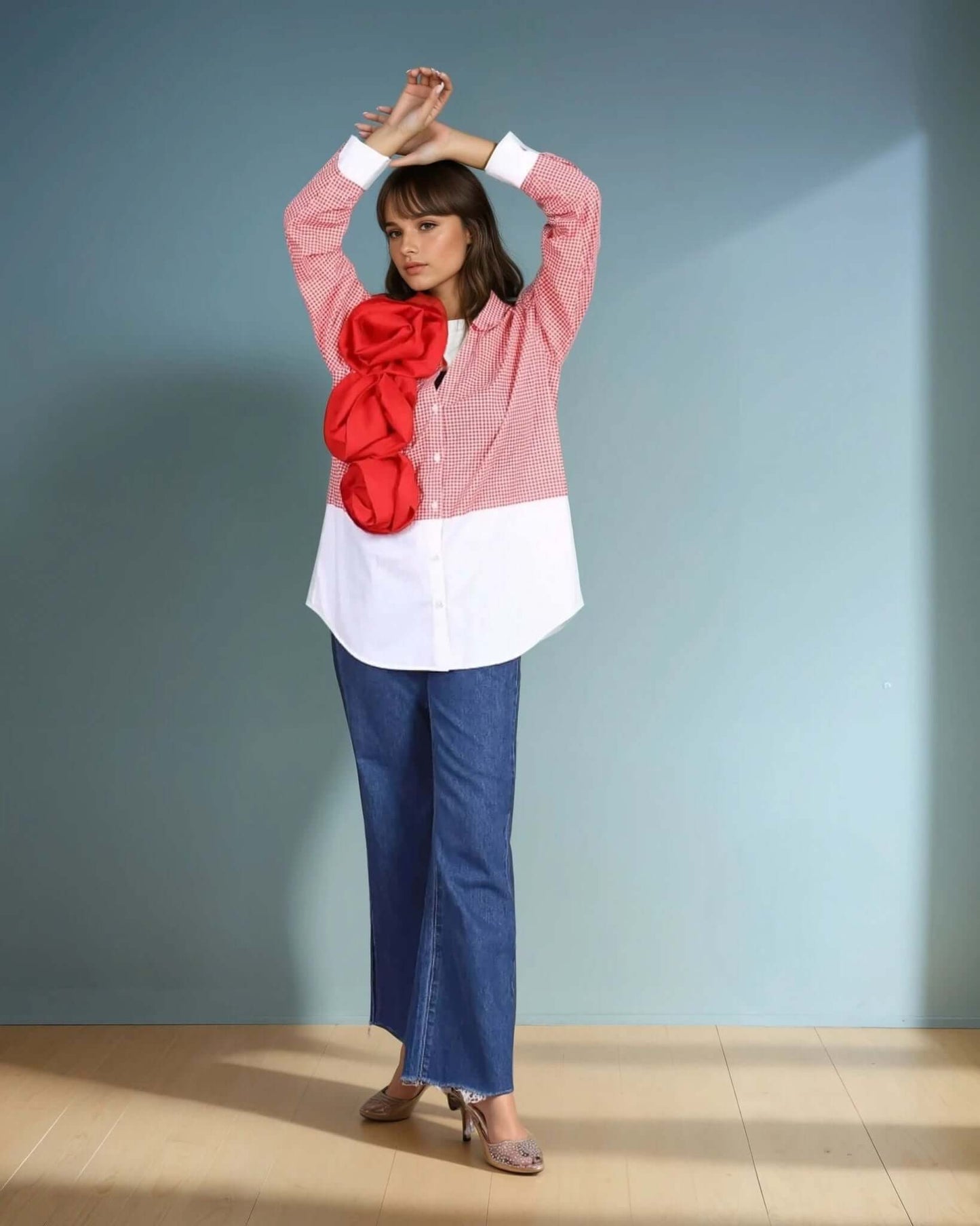 A model poses in a red and white gingham oversized shirt with a bold three-dimensional red floral applique, paired with wide-leg blue denim jeans and metallic heels. The structured yet playful design makes this shirt a standout statement piece, perfect for contemporary and fashion-forward styling.