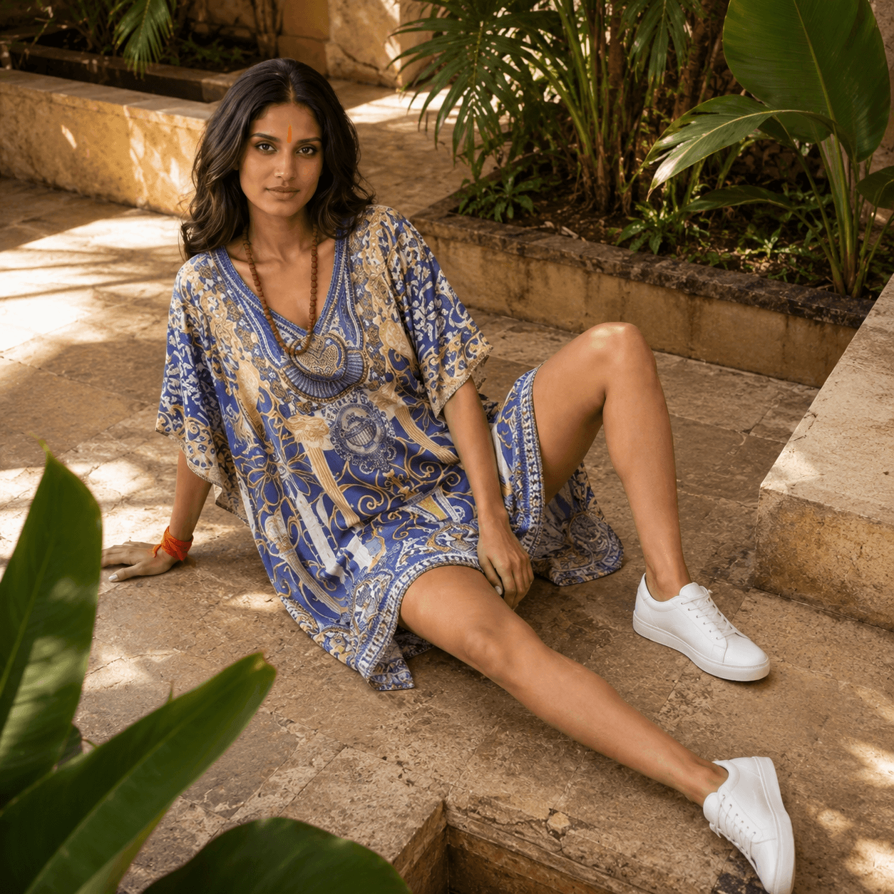 Woman in a house of majisha blue patterned kaftan dress sitting on stone steps with plants around