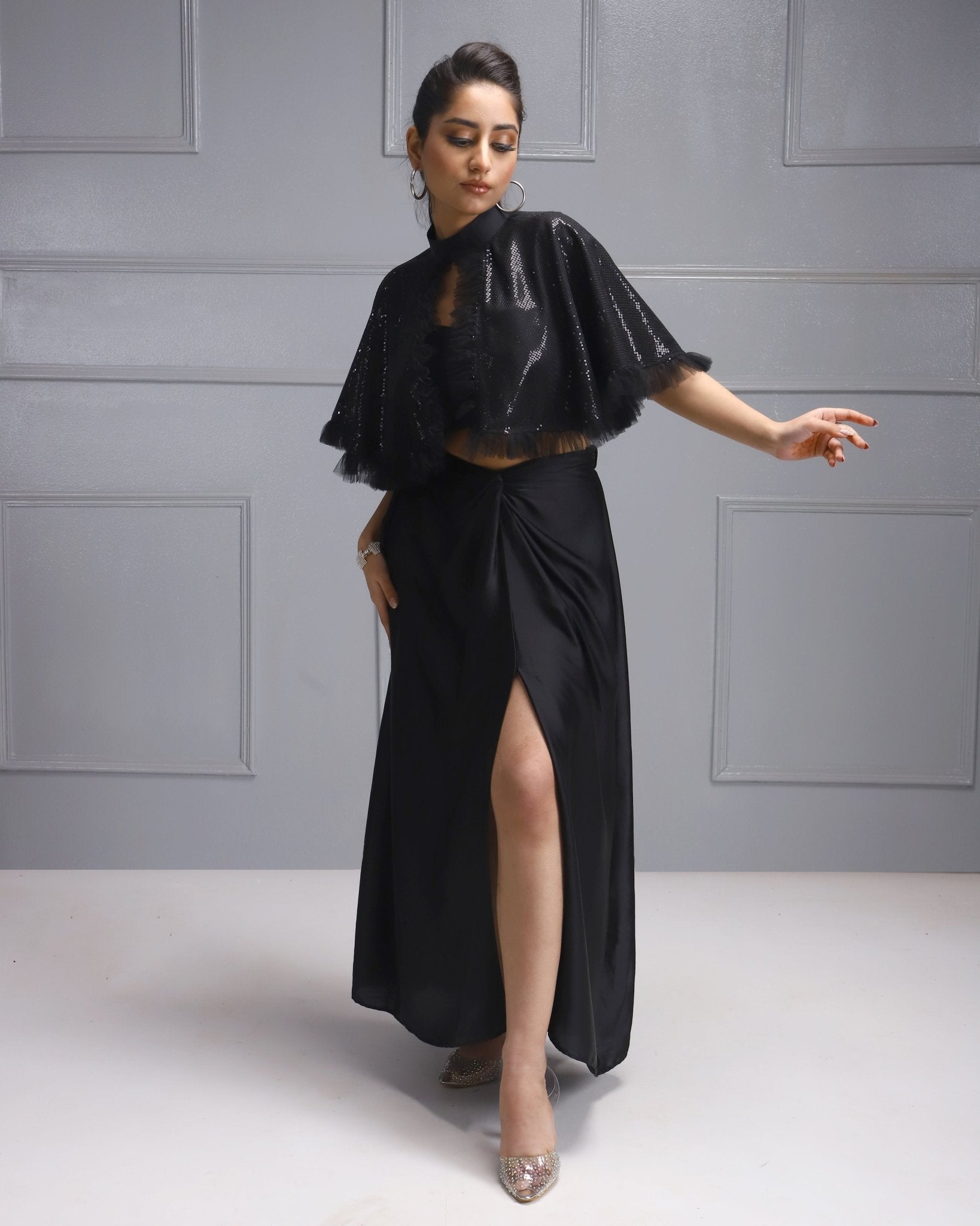 A woman wearing a House Of Majisha black outfit with a flowing slit skirt and sequined cape, striking a dynamic pose.