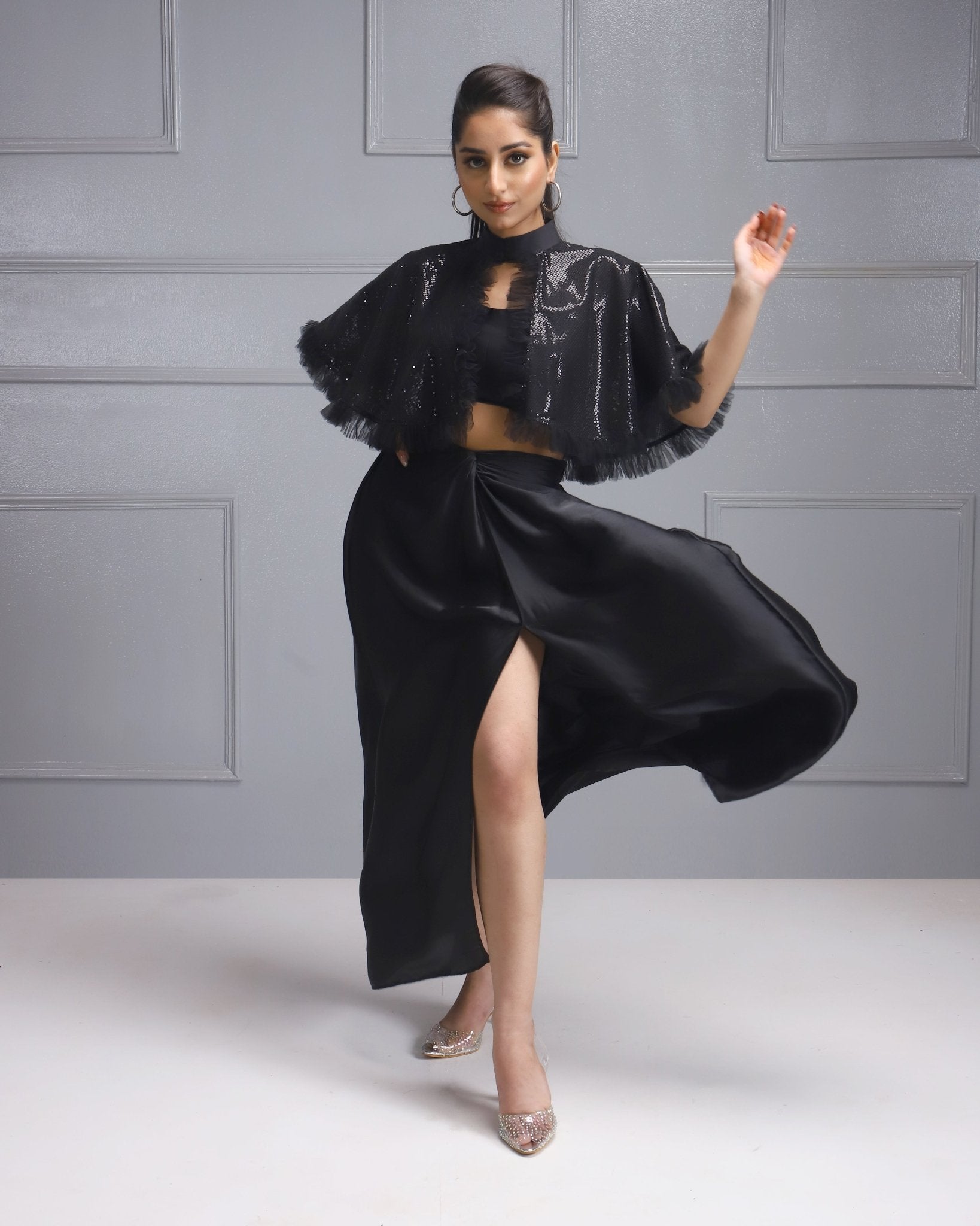 Woman in a black sequined cape and slit skirt from House Of Majisha, posing elegantly with one leg extended.
