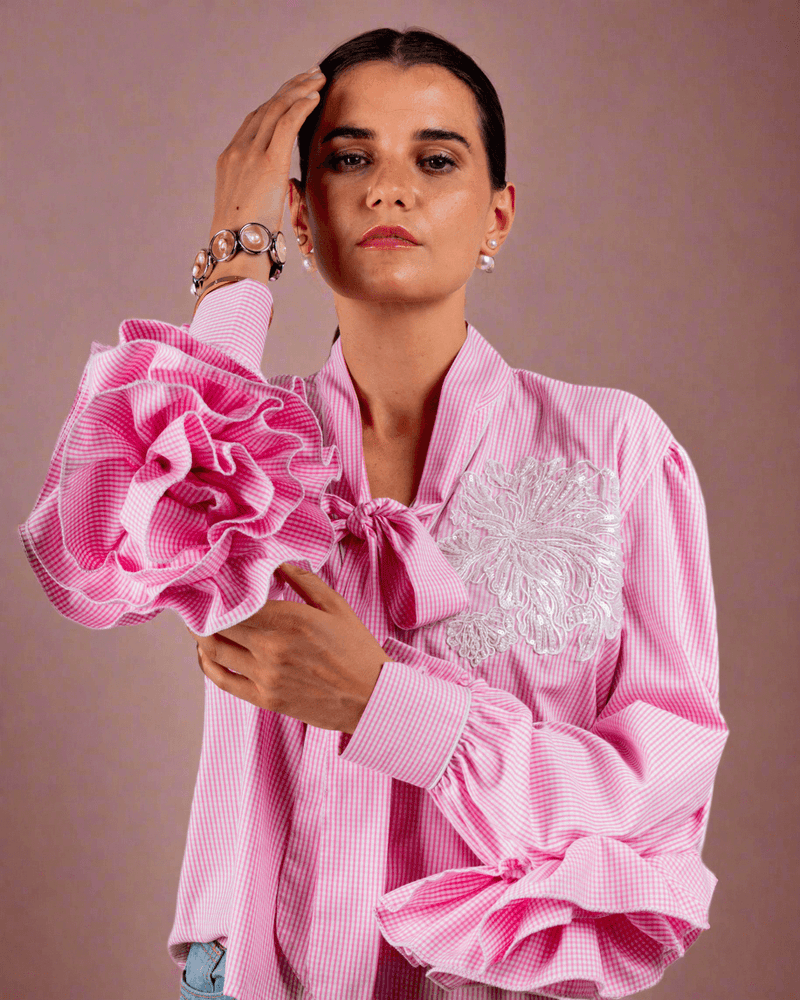 Woman wearing pink cotton shirt with ruffle sleeves, tie collar, and intricate embroidery, paired with blue jeans with metallic eyelet details