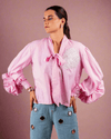 Woman wearing pink cotton shirt with ruffle sleeves, tie collar, and intricate embroidery, paired with blue jeans with metallic eyelet details