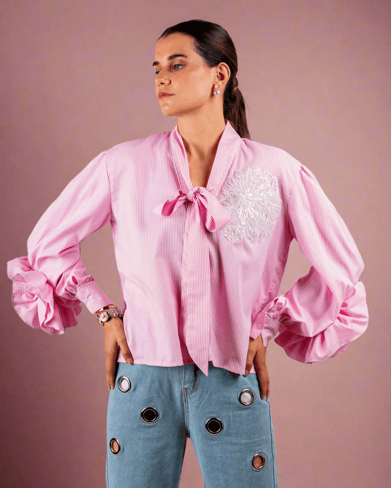 Woman wearing pink cotton shirt with ruffle sleeves, tie collar, and intricate embroidery, paired with blue jeans with metallic eyelet details