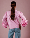 Woman wearing pink cotton shirt with ruffle sleeves, tie collar, and intricate embroidery, paired with blue jeans with metallic eyelet details