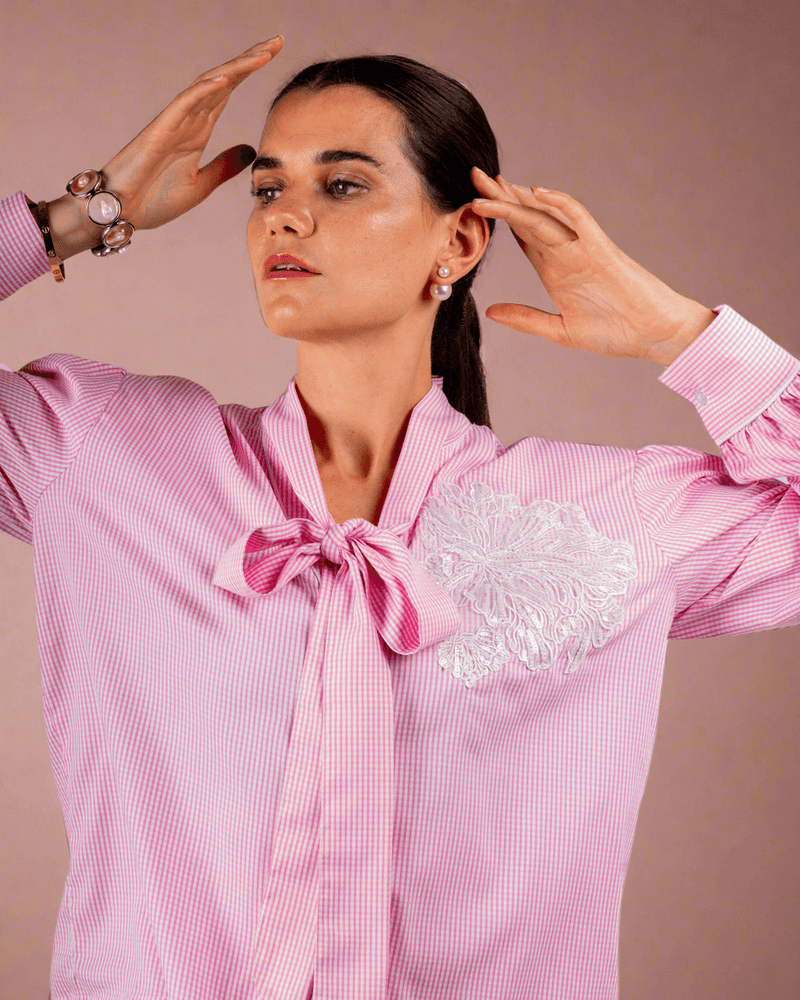 Woman wearing pink cotton shirt with ruffle sleeves, tie collar, and intricate embroidery, paired with blue jeans with metallic eyelet details