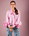 Woman wearing pink cotton shirt with ruffle sleeves, tie collar, and intricate embroidery, paired with blue jeans with metallic eyelet details