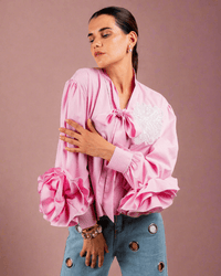 Woman wearing pink cotton shirt with ruffle sleeves, tie collar, and intricate embroidery, paired with blue jeans with metallic eyelet details