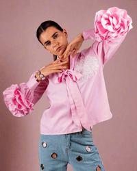 Woman wearing pink cotton shirt with ruffle sleeves, tie collar, and intricate embroidery, paired with blue jeans with metallic eyelet details