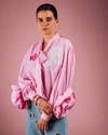 Woman wearing pink cotton shirt with ruffle sleeves, tie collar, and intricate embroidery, paired with blue jeans with metallic eyelet details