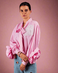 Woman wearing pink cotton shirt with ruffle sleeves, tie collar, and intricate embroidery, paired with blue jeans with metallic eyelet details