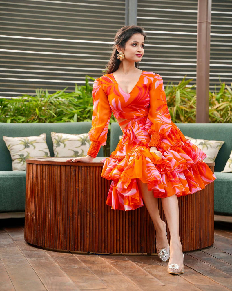 A model showcasing the Starlit Elegance Coord Dress by House Of Majisha, featuring a vibrant orange floral print with a flattering V-neck and full sleeves, perfect for brunch outings or picnics. The dress is knee-length with playful ruffles and a stylish cutout at the waist, embodying a chic skater style.