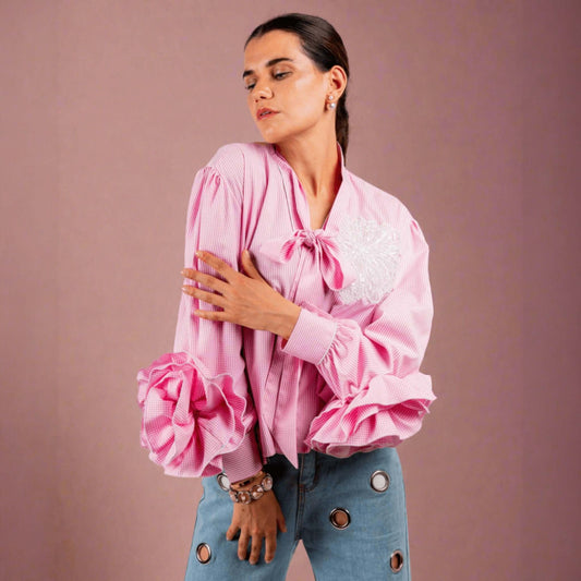 Woman wearing pink cotton shirt with ruffle sleeves, tie collar, and intricate embroidery, paired with blue jeans with metallic eyelet details