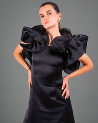 A woman in a House Of Majisha black dress with oversized ruffles, striking a strong pose with arms crossed.