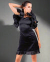 Woman posing in a black satin dress by House Of Majisha, featuring bold ruffled details at the shoulder and hem.