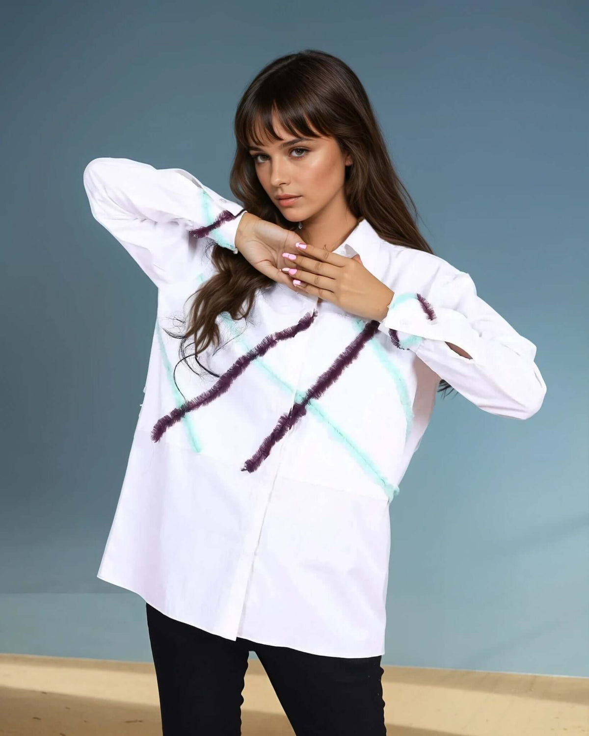 Model wearing a white oversized button-down shirt with abstract diagonal brushstroke details in purple and teal. The shirt features a relaxed fit, long sleeves with frayed accents, and is paired with sleek black pants and pointed-toe heels, creating a bold and modern look