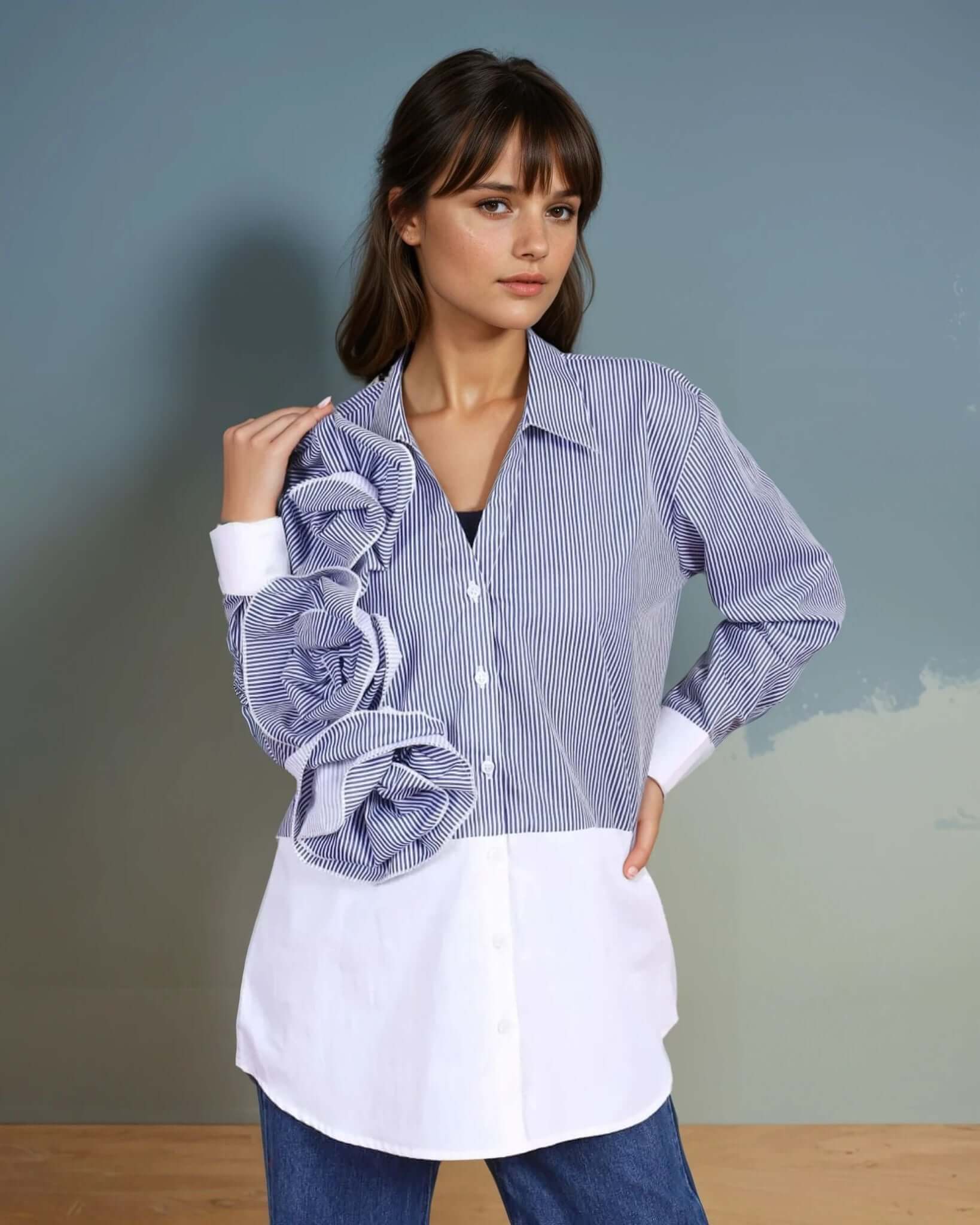 A model wearing a blue and white striped oversized button-down shirt with dramatic three-dimensional ruffle detailing on one side, paired with wide-leg denim jeans and metallic pointed-toe heels. The outfit blends structured elegance with contemporary fashion, perfect for statement styling.