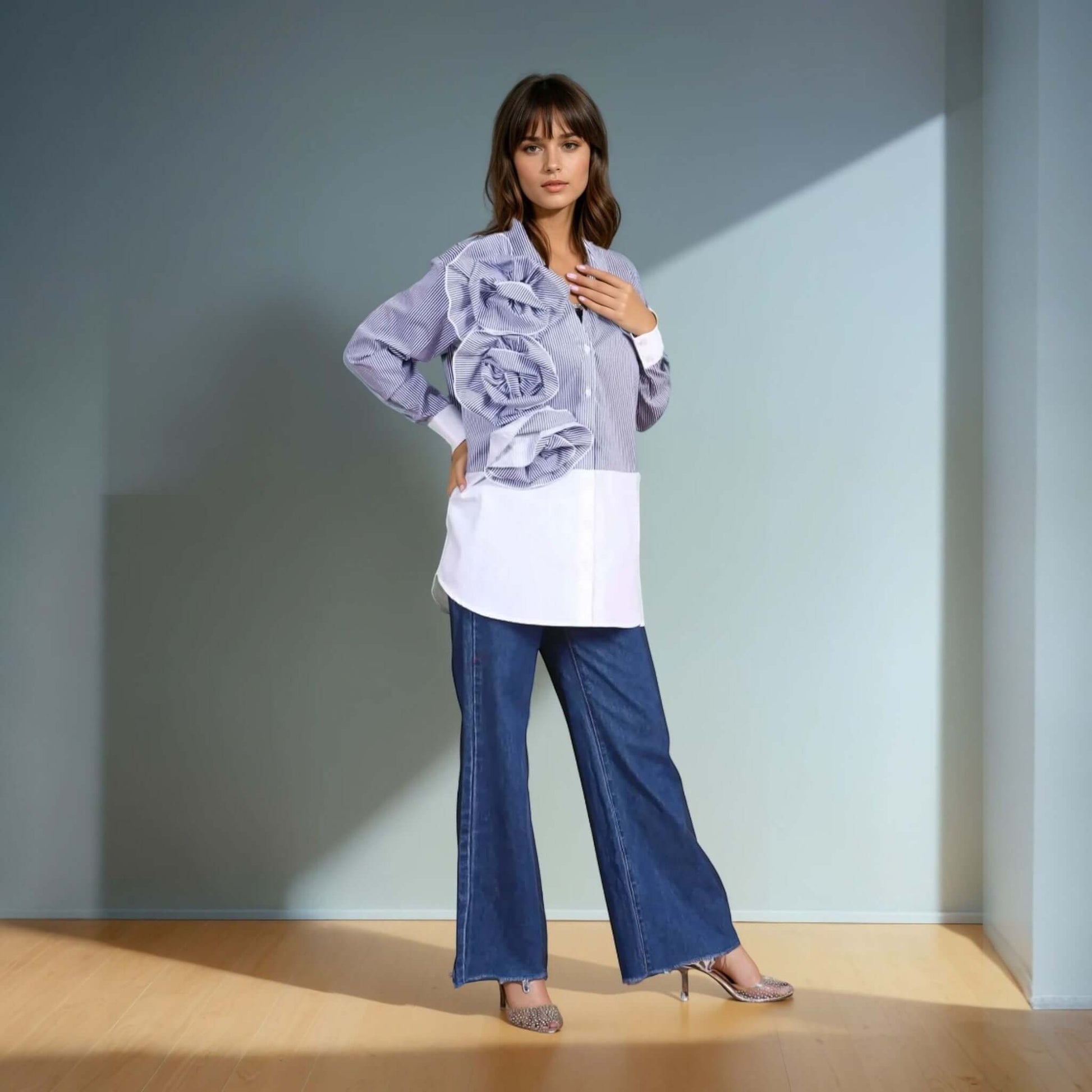 A model wearing a blue and white striped oversized button-down shirt with dramatic three-dimensional ruffle detailing on one side, paired with wide-leg denim jeans and metallic pointed-toe heels. The outfit blends structured elegance with contemporary fashion, perfect for statement styling.