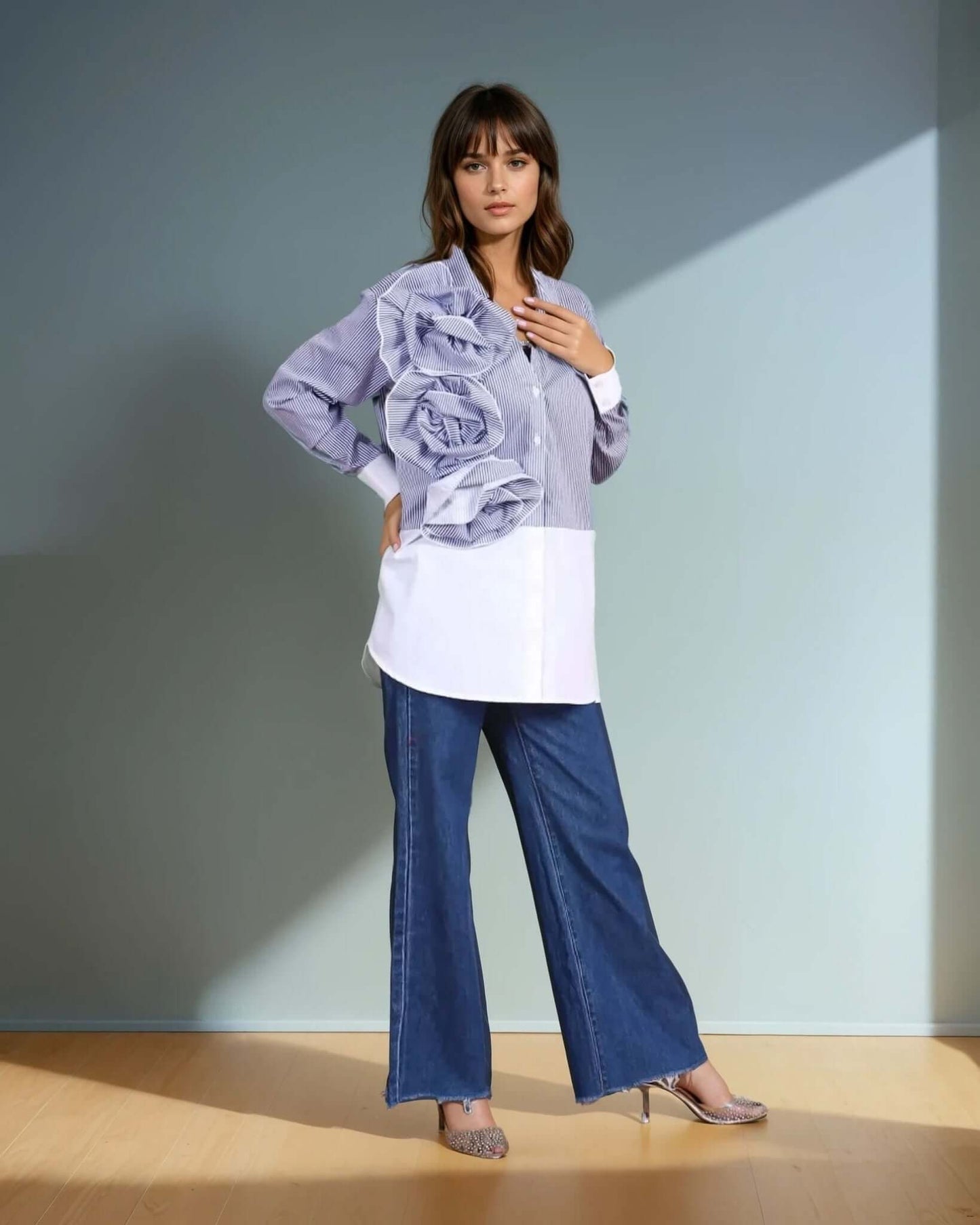 A model wearing a blue and white striped oversized button-down shirt with dramatic three-dimensional ruffle detailing on one side from House Of Majisha, paired with wide-leg denim jeans and metallic pointed-toe heels. The outfit blends structured elegance with contemporary fashion, perfect for statement styling.