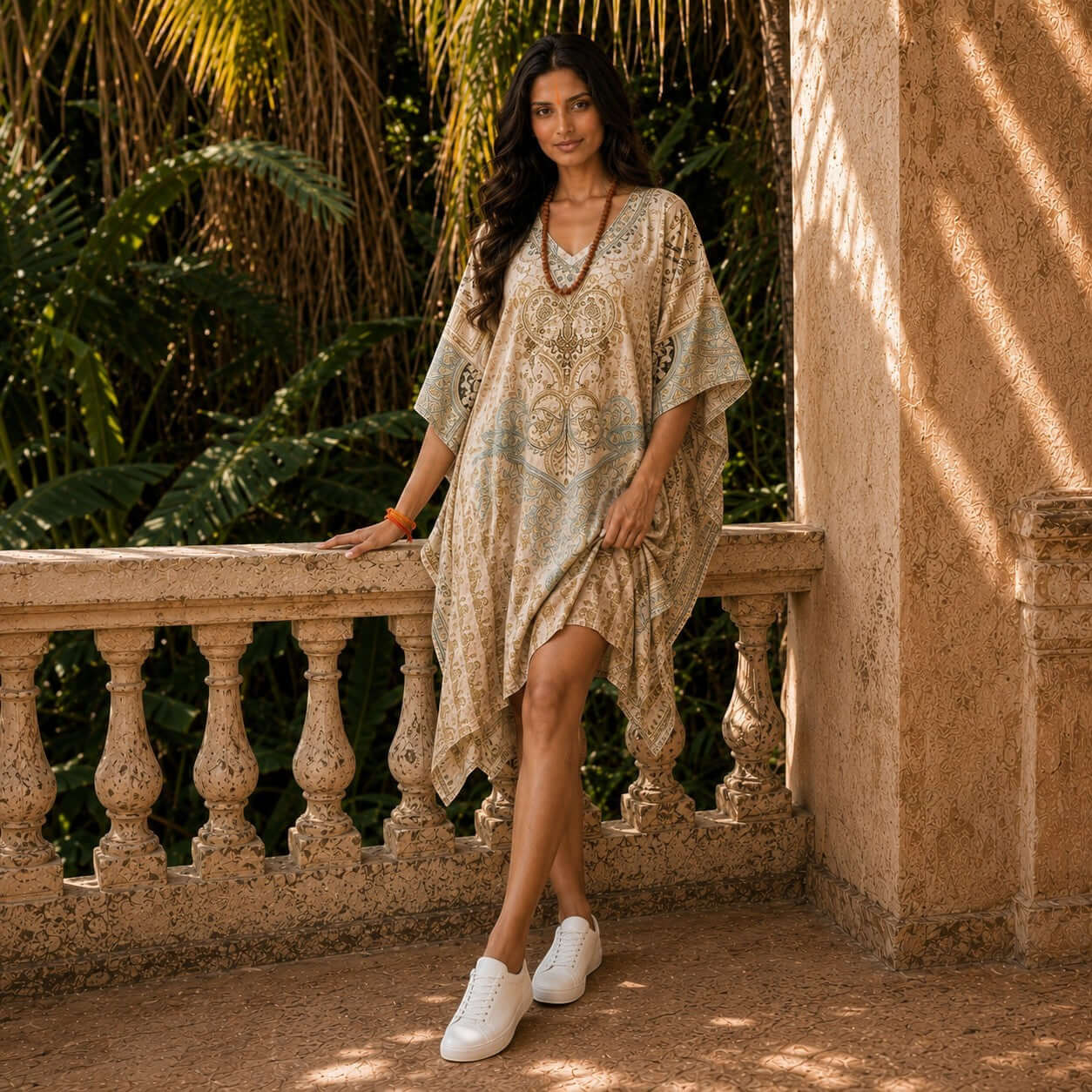 Woman in a house of majisha patterned kaftan dress standing on a stone balcony with greenery in the background.