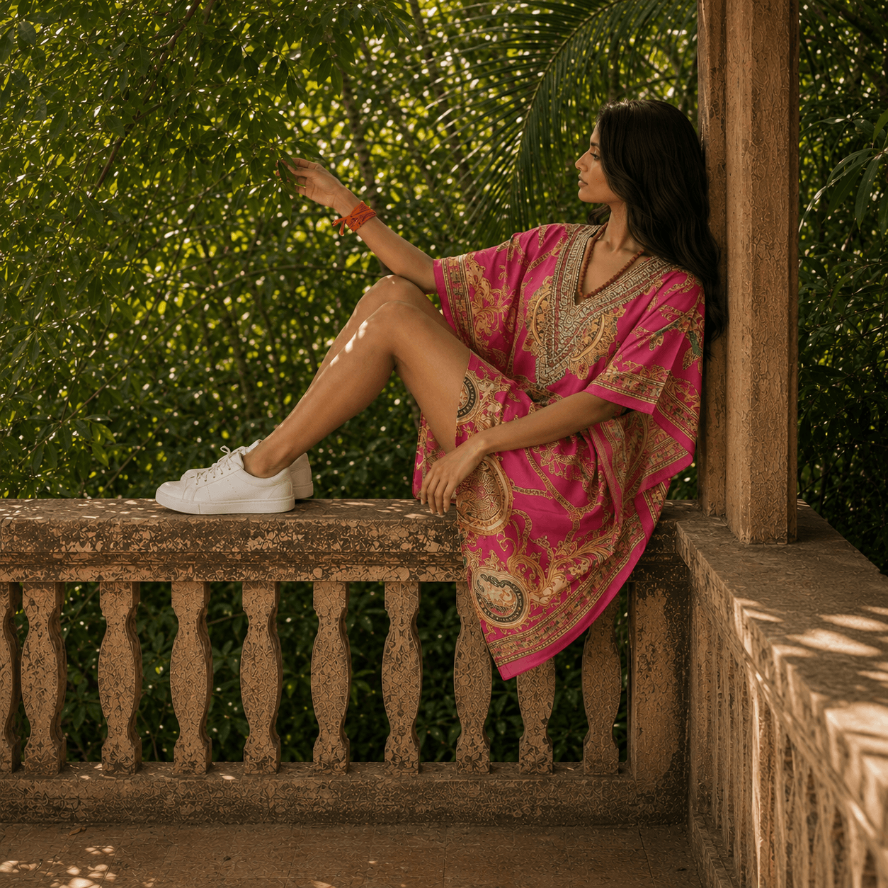 Woman in a pink dress with gold patterns kaftan from house of majisha  sitting on a stone railing outdoors.