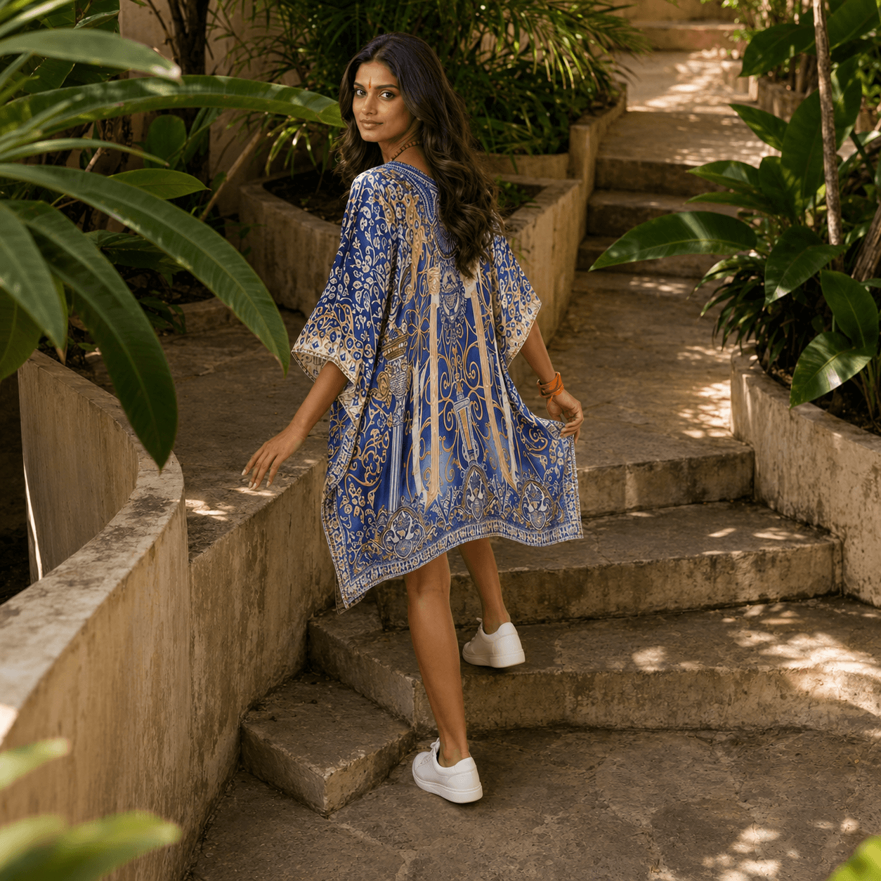 Woman in a house of majisha blue patterned kaftan dress standing on stone steps surrounded by greenery
