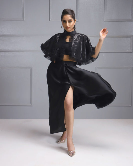 Woman in a black sequined cape and slit skirt from House Of Majisha, posing elegantly with one leg extended.